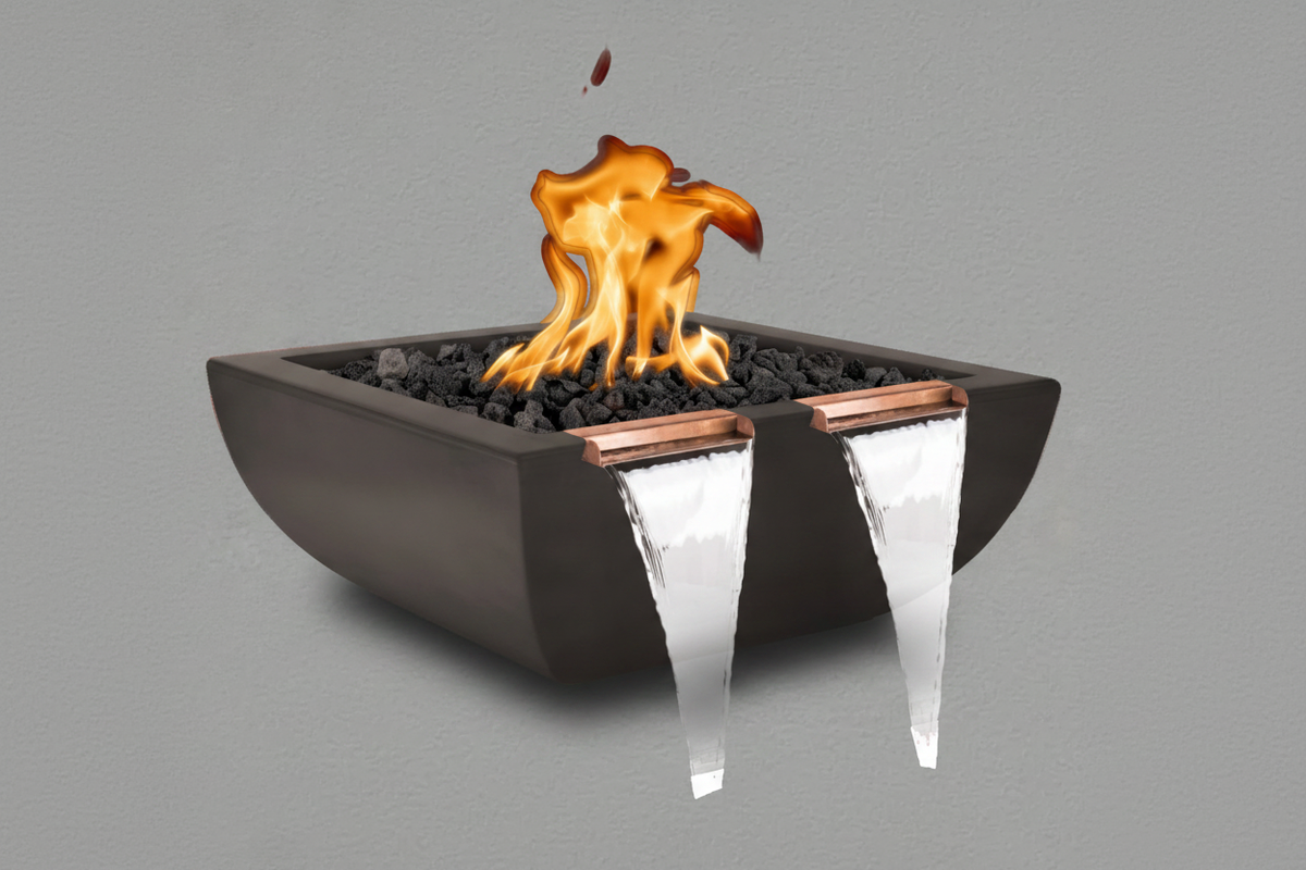 A modern outdoor fire pit bowl with a matte black finish, filled with black river rocks and featuring two copper spout details with clear waterfall-like water flowing from the front, set against a solid gray background.