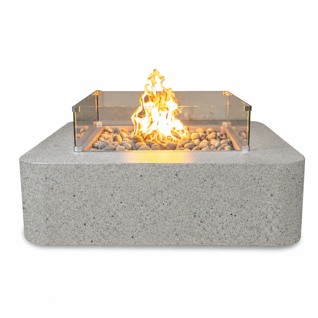A modern square fire pit with a textured gray stone-look base, clear glass wind guard, and glowing orange-yellow flames over decorative river rocks, shown on a solid white background.