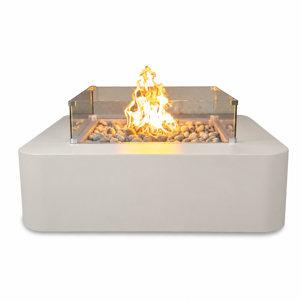 A modern outdoor fire pit table with a matte light gray concrete base, a clear glass wind guard, and a bed of gray river rocks with a bright orange and yellow flame burning inside, shown on a solid white background.