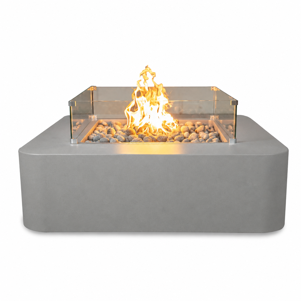 A modern square outdoor fire pit with a matte gray concrete-look base, clear glass wind guard, and glowing orange-yellow flames over decorative river rocks, shown on a plain white background.