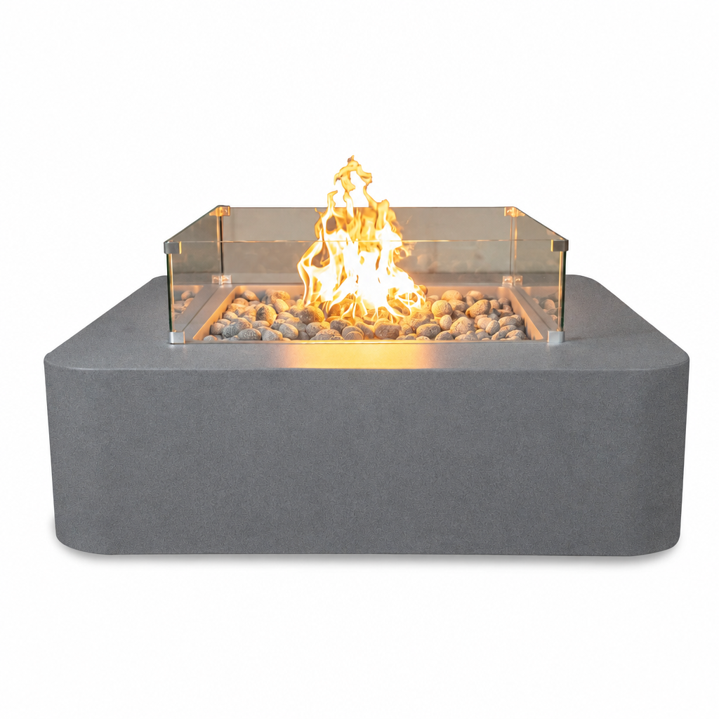 A modern square outdoor fire pit with a matte gray concrete-look base, clear glass sides, and glowing orange-yellow flames over light-colored stones, shown on a plain white background.