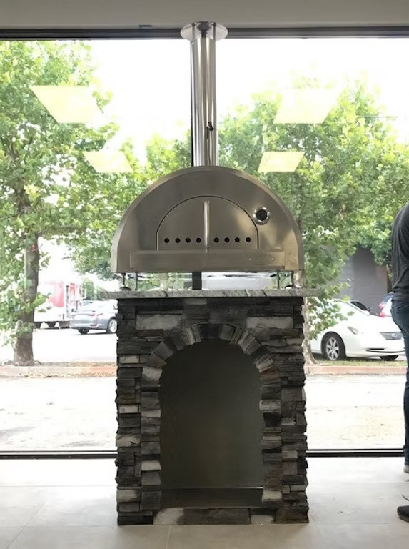 Outdoor pizza oven on a stone base in front of a glass door with a view of trees and cars outside.