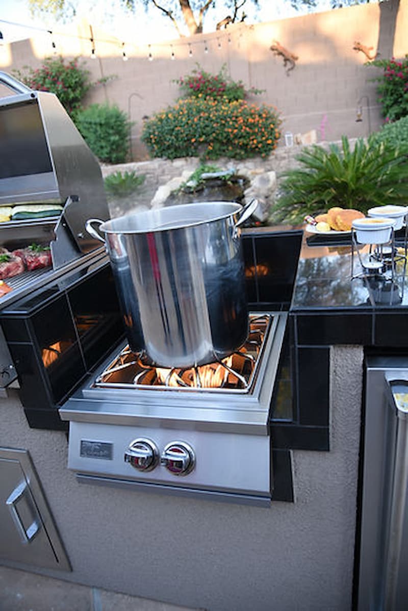 Outdoor kitchen with a grill and pot on a stove, surrounded by garden elements.