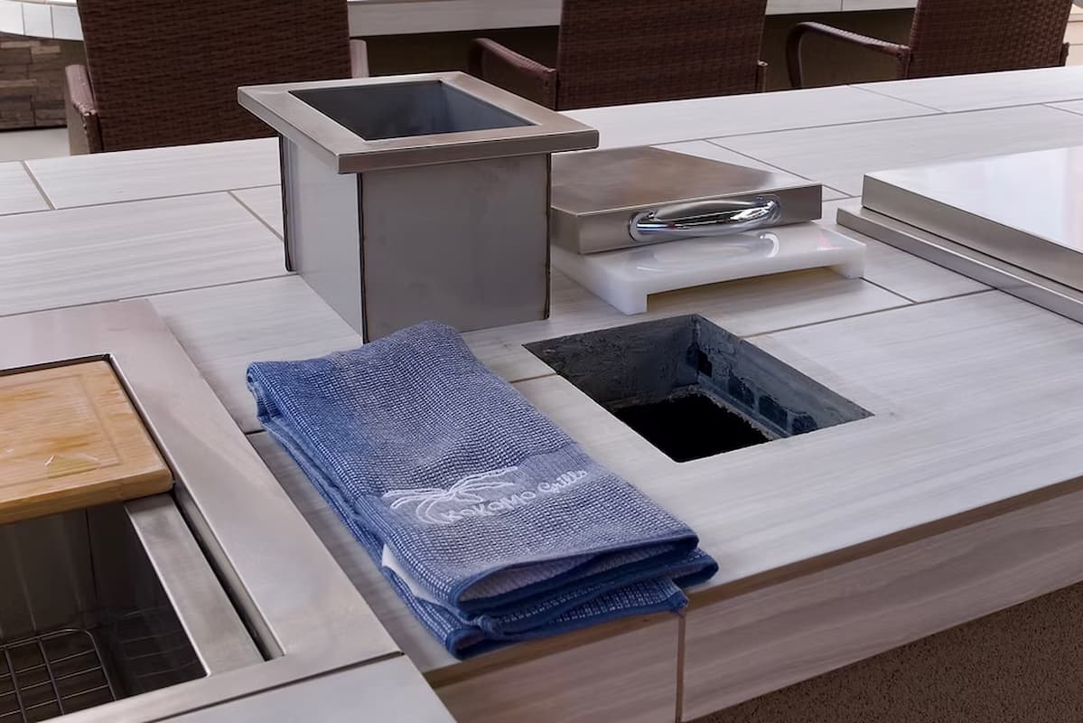 Outdoor kitchen island detail showing a folded blue towel with white script, stainless steel cookware, and light wood surfaces.
