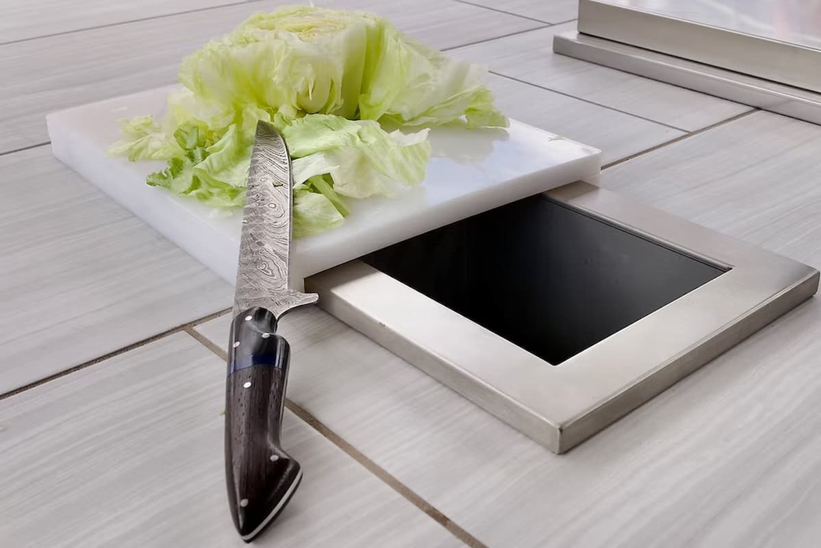 A stainless steel pull-out knife block and drawer base shown open on a light wood floor, with a white cutting board, a knife, and lettuce placed above the recessed compartment.