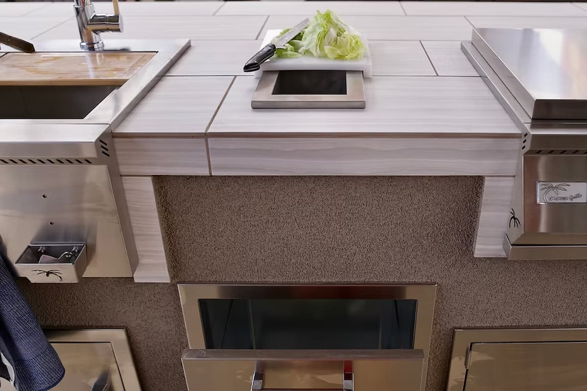 A close-up of a modern outdoor kitchen island featuring light wood-look tile countertops, stainless steel built-in appliances, a textured beige center panel, and a small cutting board with lettuce on top.