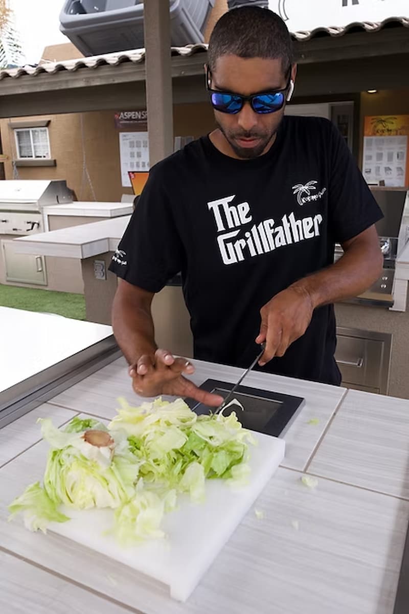 Man preparing food outdoors, wearing a black 'The Grillfather' t-shirt.