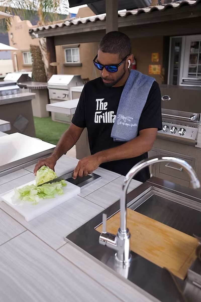 A man wearing a black t-shirt with 'The Grillf*ck' printed on it, blue sunglasses, and a gray towel over his shoulder, slicing lettuce on a white cutting board at an outdoor kitchen.