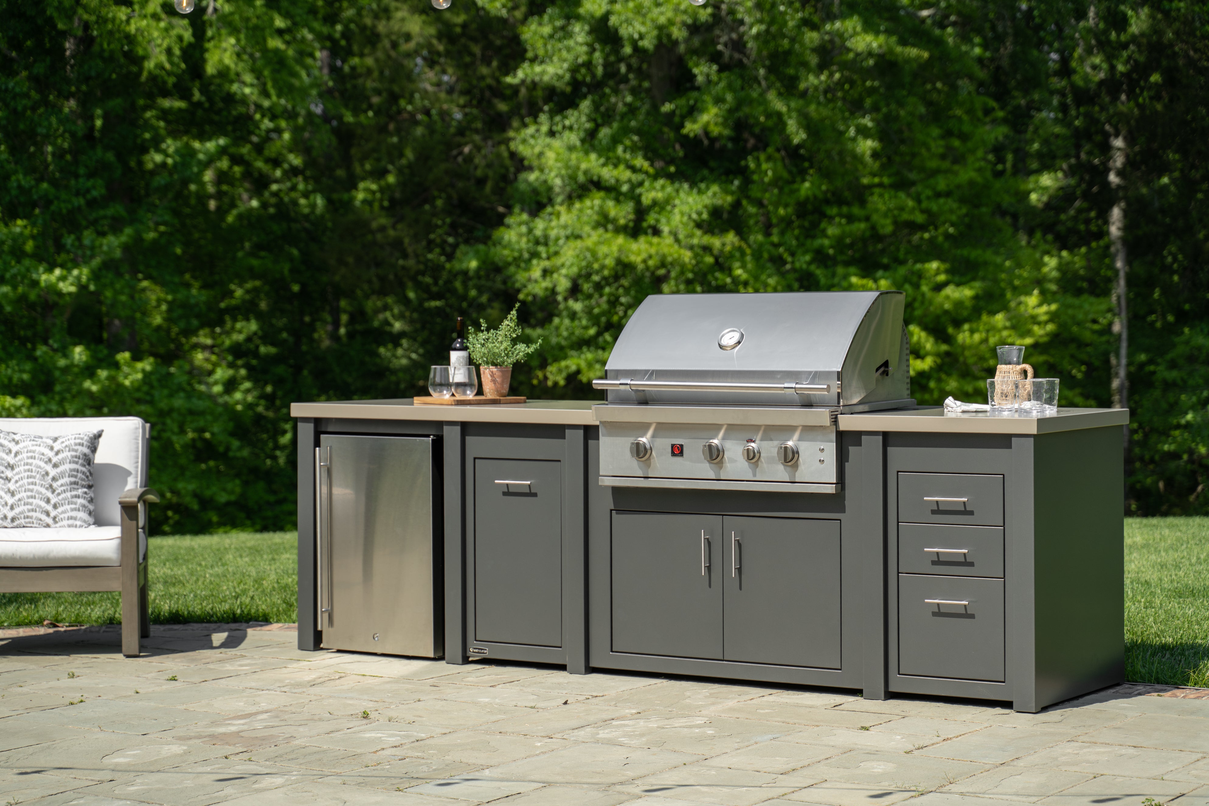 An outdoor kitchen island set with a stainless steel gas grill, a refrigerator, and gray cabinets on a stone patio with a lush green forest background.