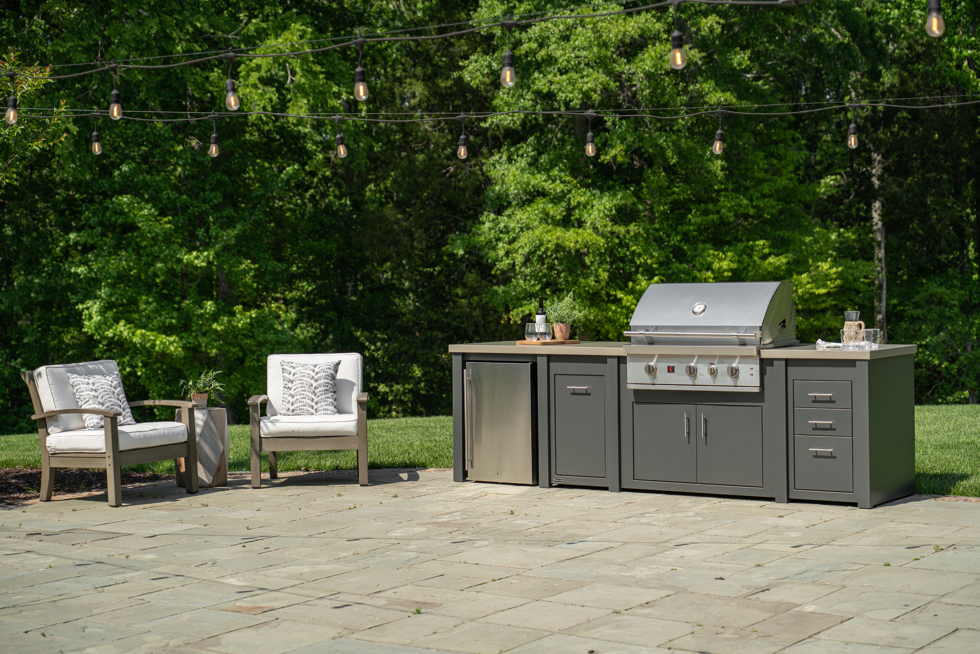 An outdoor kitchen island set with a stainless steel gas grill, gray cabinets, and a stone patio in front of a lush green forest backdrop with hanging string lights.