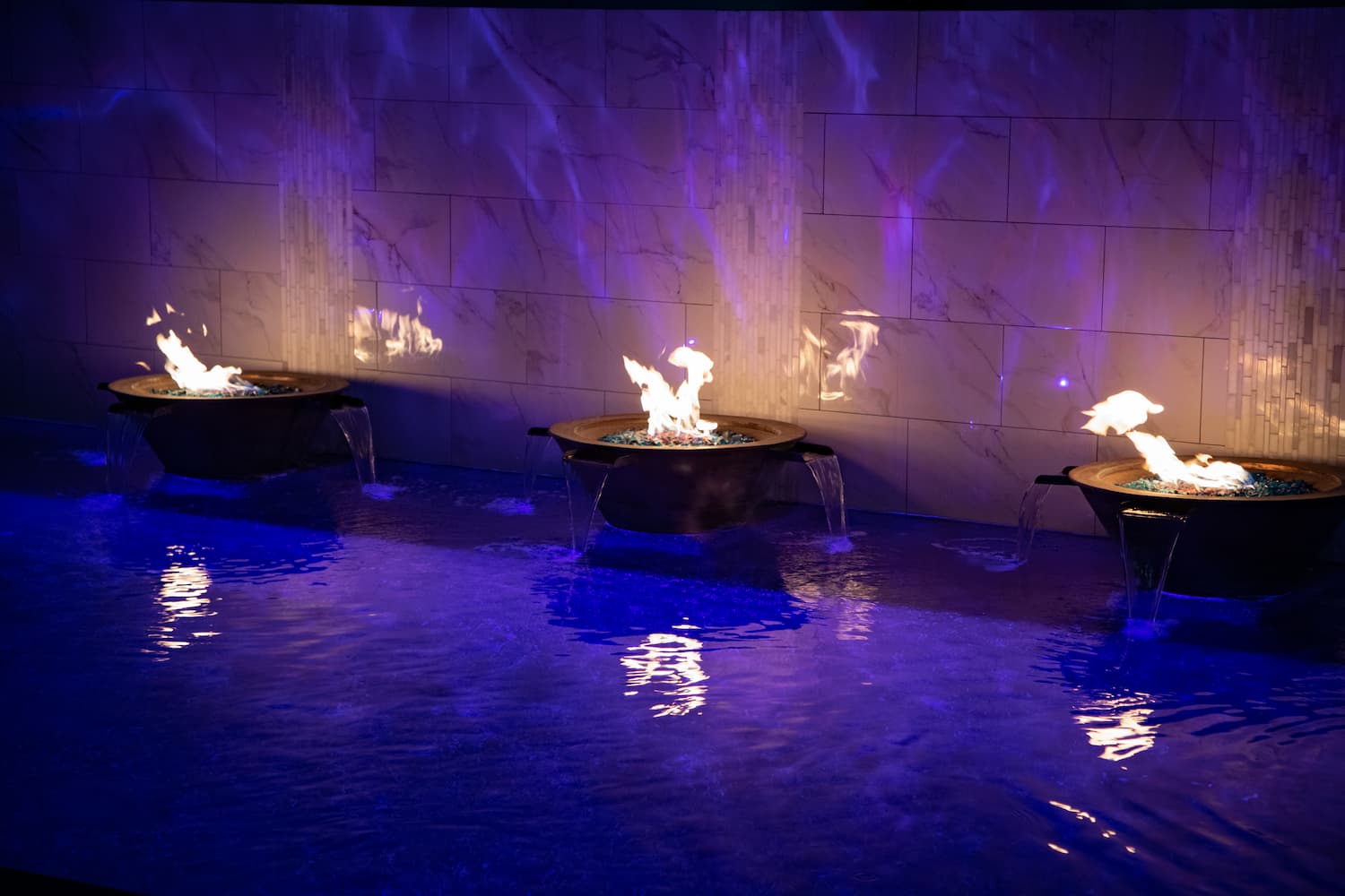 Three outdoor fire pits with active flames, set in a shallow water feature against a tiled wall illuminated by purple lighting.