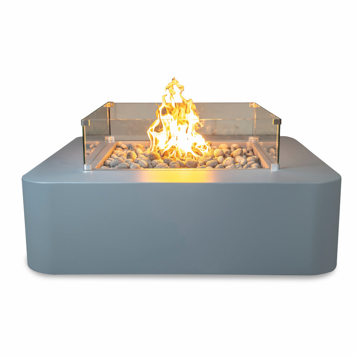 A modern square outdoor fire pit with a matte gray base, clear glass wind guard, and glowing orange-yellow flames over decorative river rocks, shown on a plain white background.