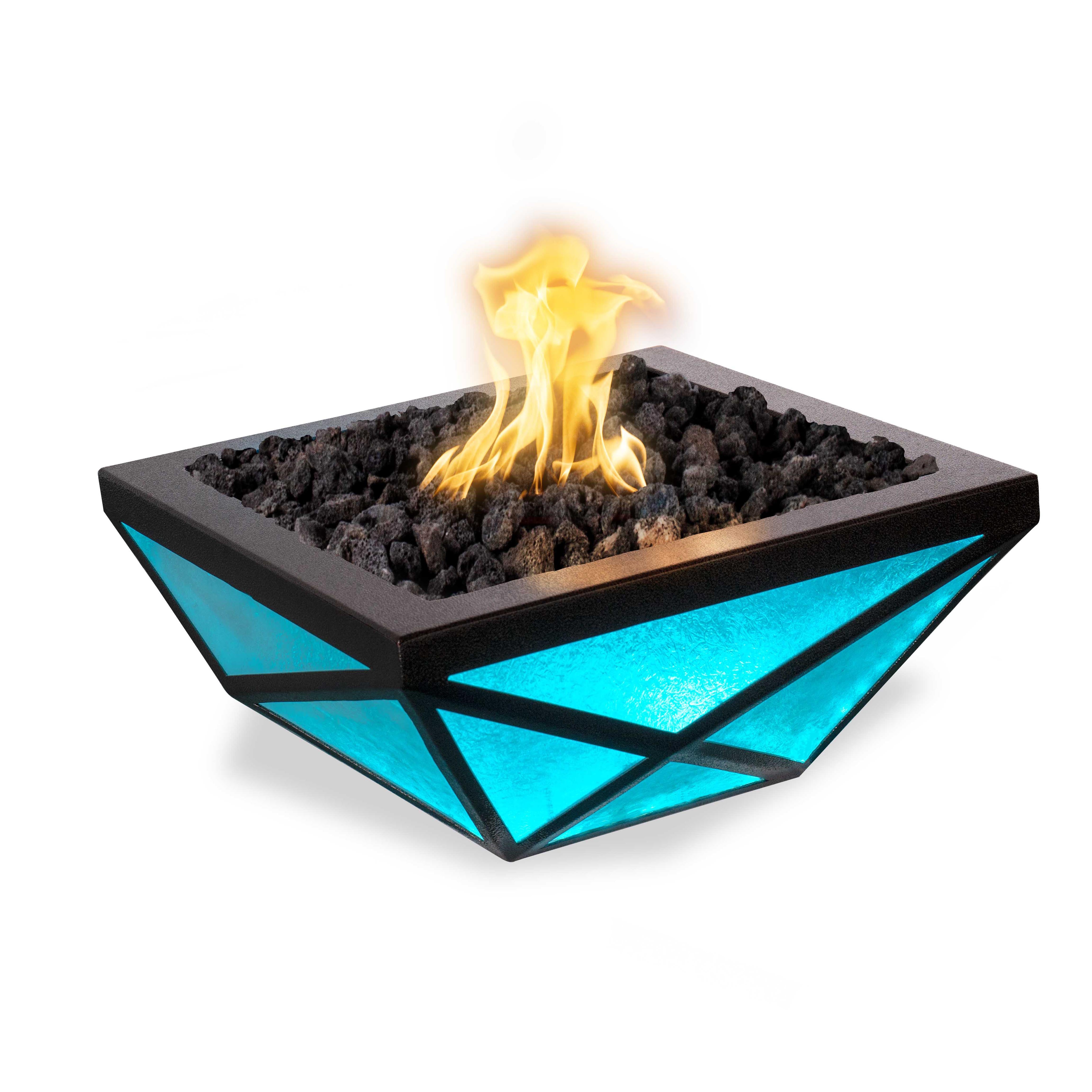 A modern square fire pit table with a black metal frame and glowing turquoise blue faceted glass panels, filled with black lava rocks and lit with a bright yellow-orange flame, isolated on a white background.
