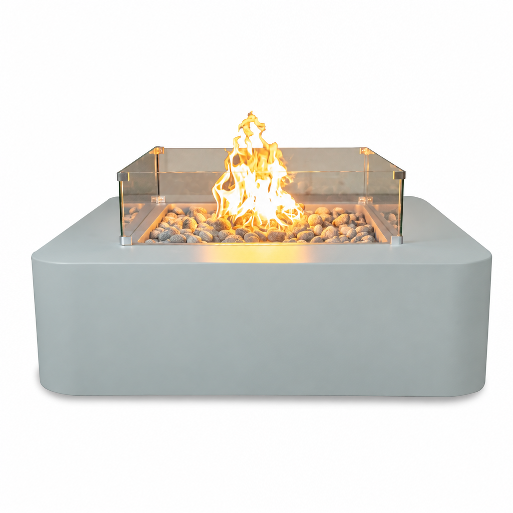 A modern square outdoor fire pit with a matte light gray base, clear acrylic wind guard, and glowing orange-yellow flames over decorative river rocks, shown on a plain white background.