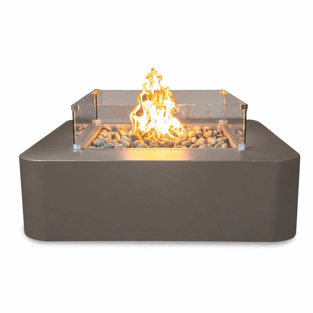 A rectangular outdoor fire pit with a matte dark gray base, clear glass wind guard, and glowing orange-yellow flames over decorative river rocks, shown on a plain white background.