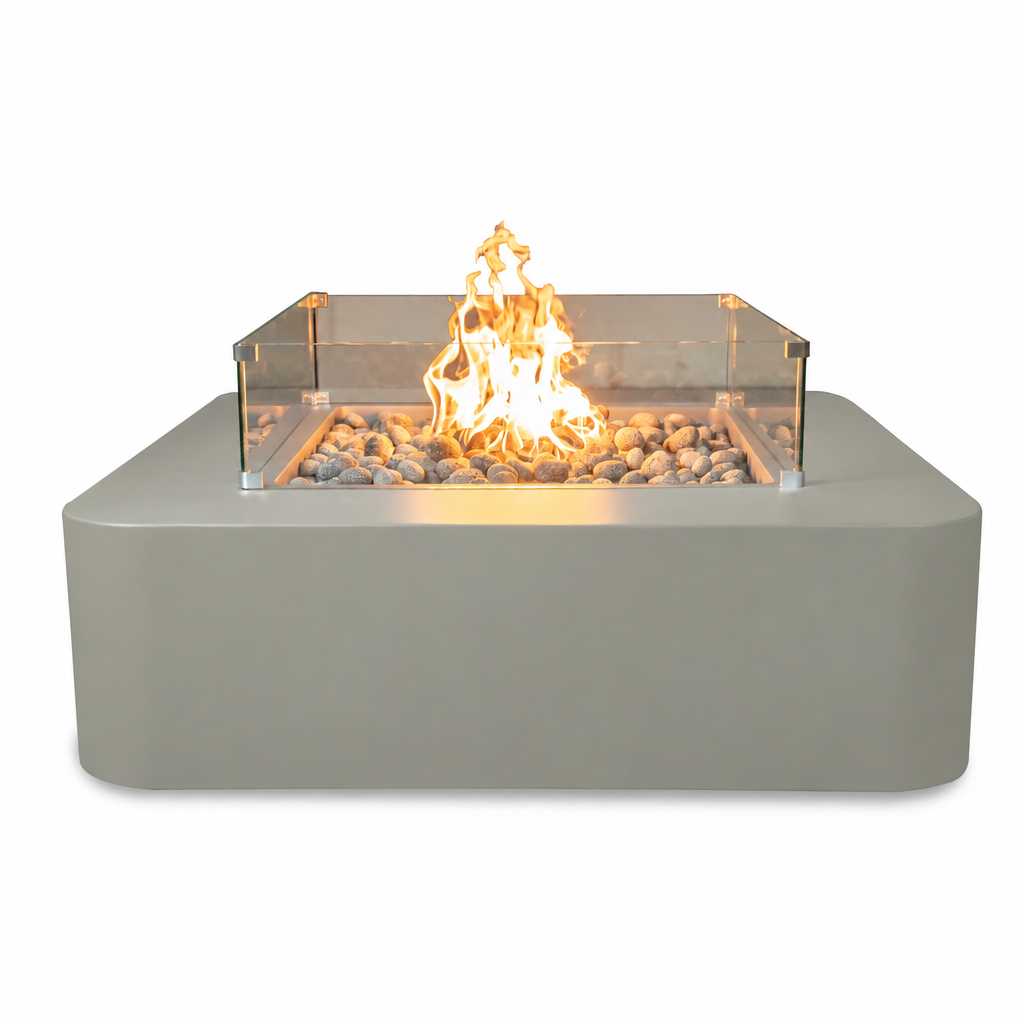 A modern square outdoor fire pit with a matte gray concrete-look base, clear glass wind guard, and glowing orange flames over decorative stones, shown on a solid white background.