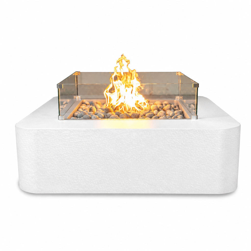 A modern outdoor fire pit with a white rectangular stone base, clear glass wind guard, and active orange-yellow flame over decorative gray stones, shown on a plain white background.