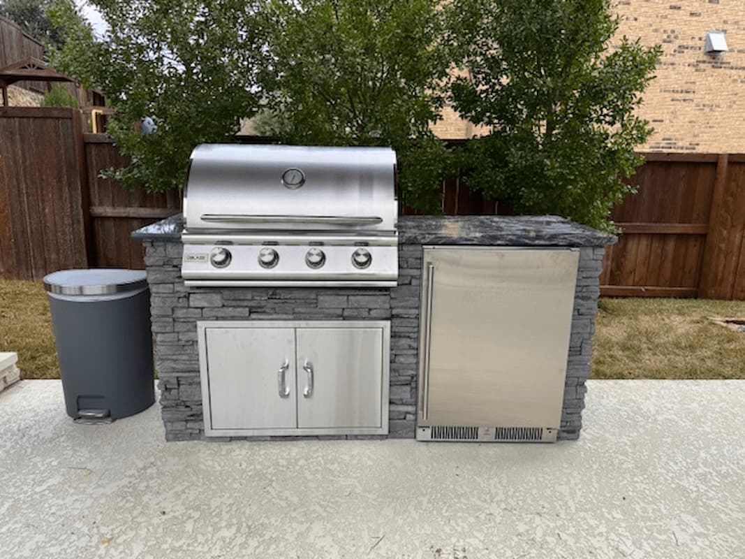 A stainless steel outdoor kitchen island set with a four-burner gas grill, stone-look cabinets, a dark gray stone countertop, and a built-in refrigerator, shown on a concrete patio in a backyard.