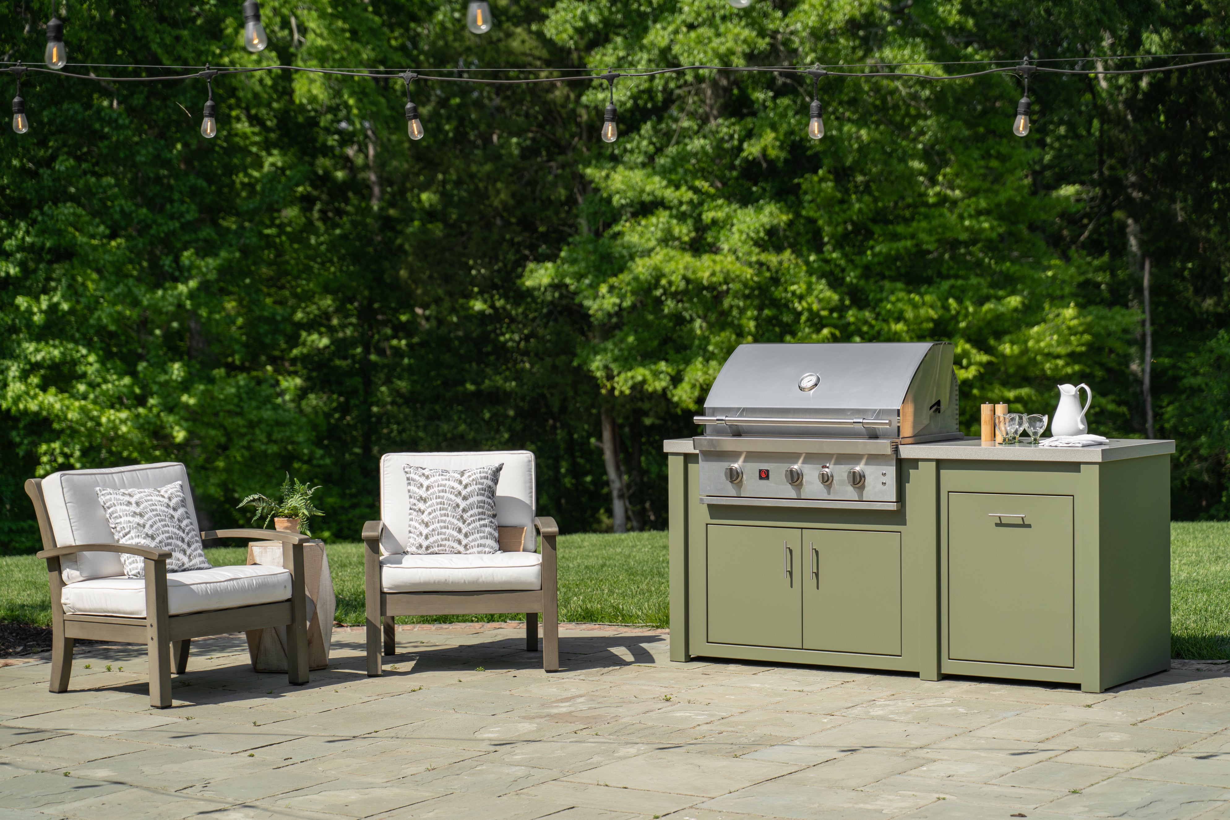 An outdoor kitchen island set featuring a stainless steel gas grill on a sage green cabinet base with a stone-look countertop, styled next to two wooden armchairs with white cushions on a stone patio in front of a lush green forest background.