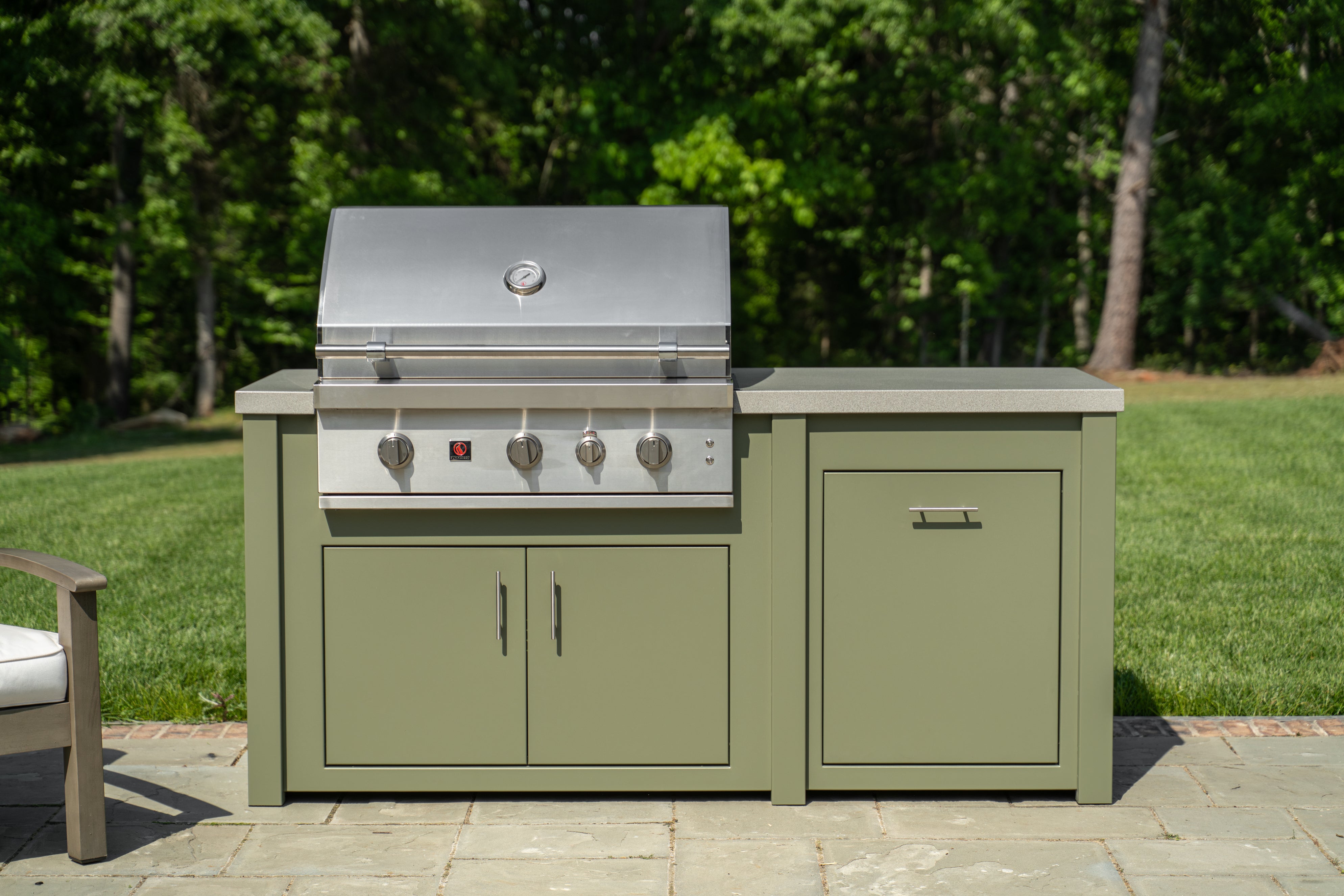 An outdoor built-in gas grill and side counter unit with sage green cabinets and a stainless steel grill top, set on a stone patio with a green lawn and trees in the background.