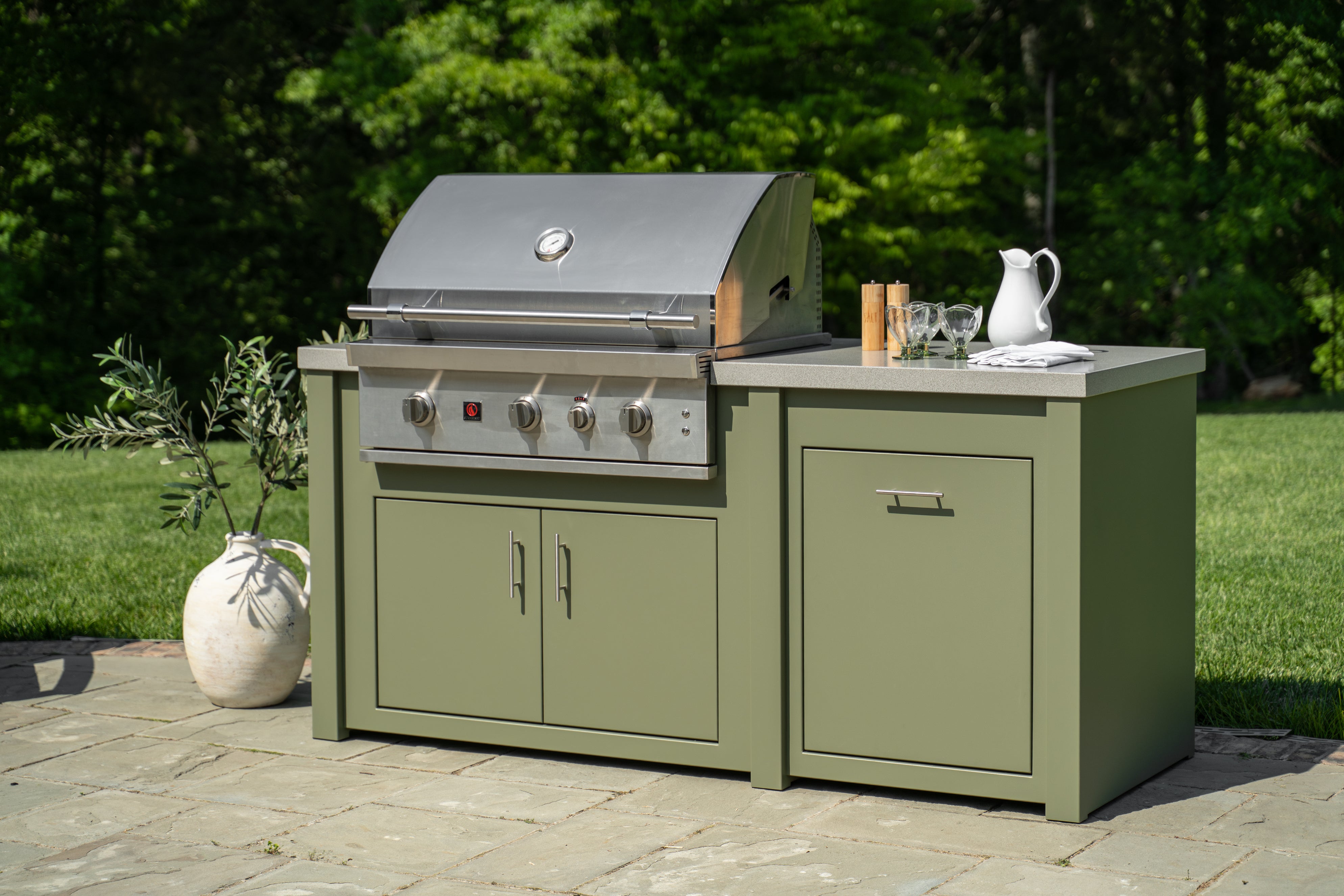 An outdoor kitchen island set featuring a stainless steel gas grill and a matching sage green cabinet base, set on a stone patio with a green lawn and trees in the background.
