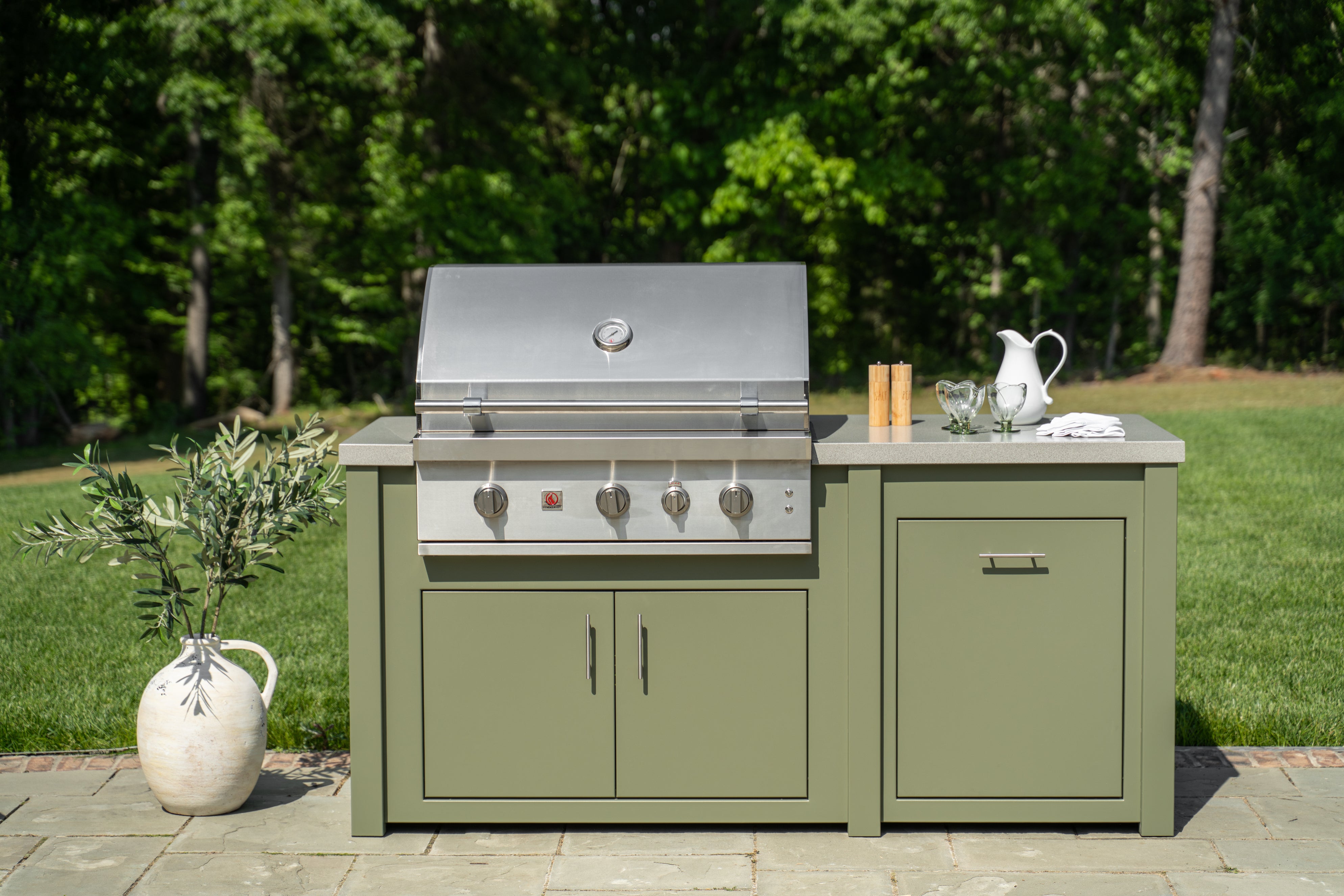 An outdoor kitchen island set featuring a stainless steel gas grill mounted on a sage green cabinet base with a stone-look countertop, shown in a backyard setting with a grassy lawn and trees in the background.