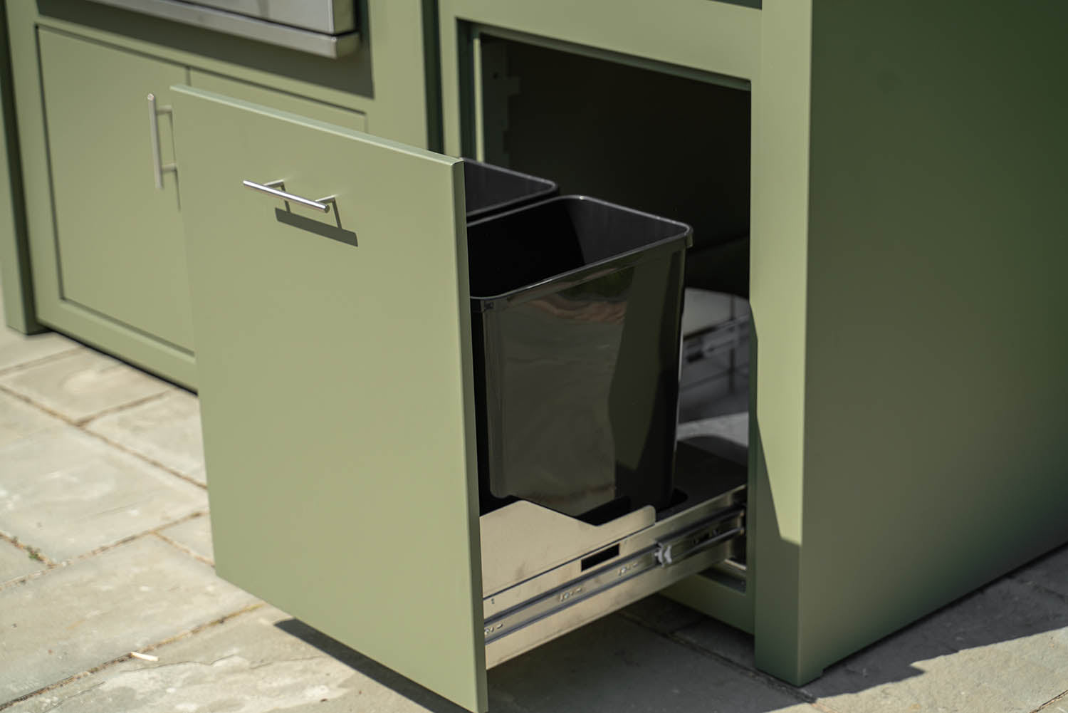 An olive green metal storage cabinet with a sliding door open, revealing a black plastic bin inside on a metal pull-out tray, set outdoors on a stone pavement.
