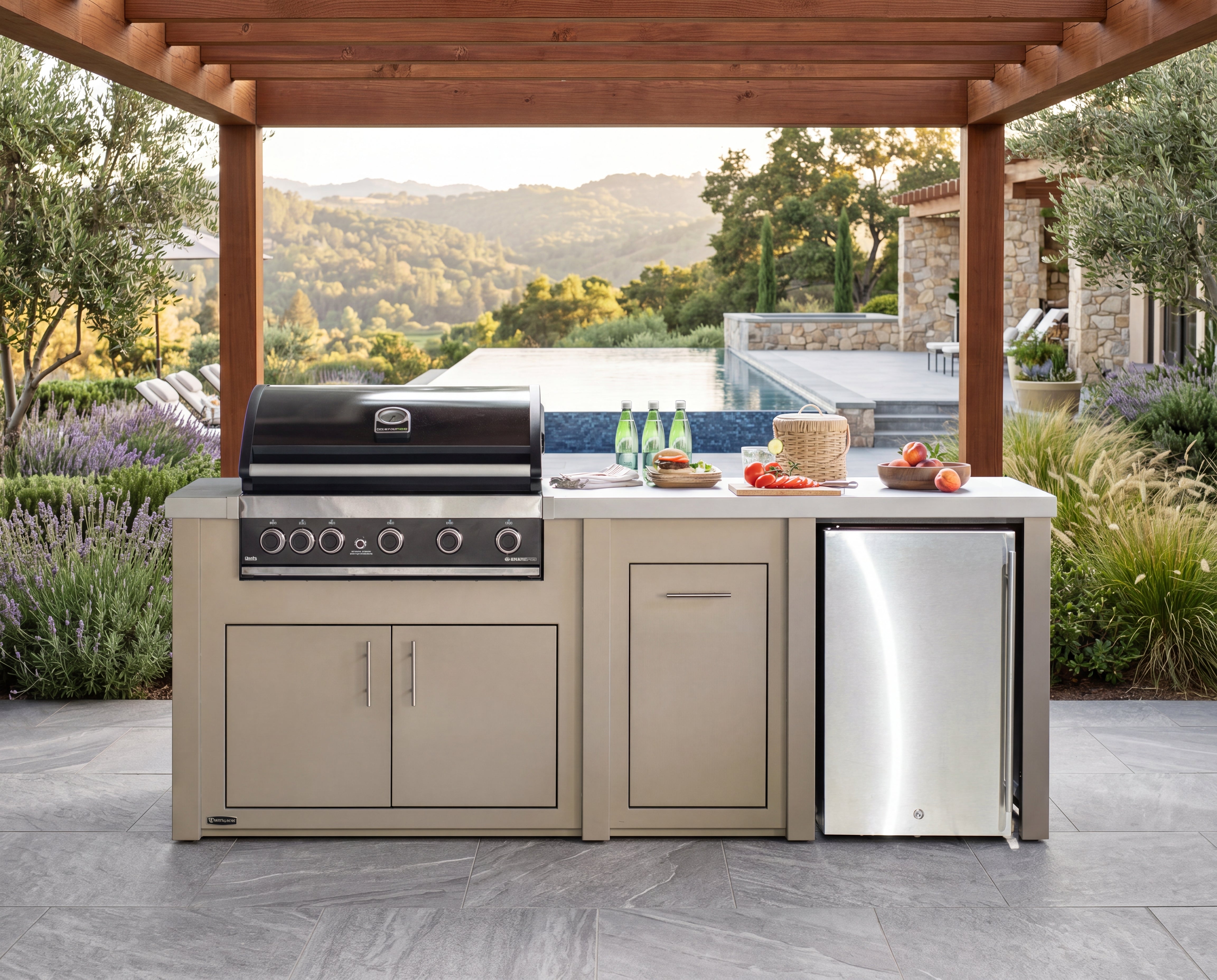 Outdoor kitchen island with a black gas grill on the left, a stainless steel mini refrigerator on the right, and a white countertop with food and bottles, set under a wooden pergola with a pool and hills in the background.