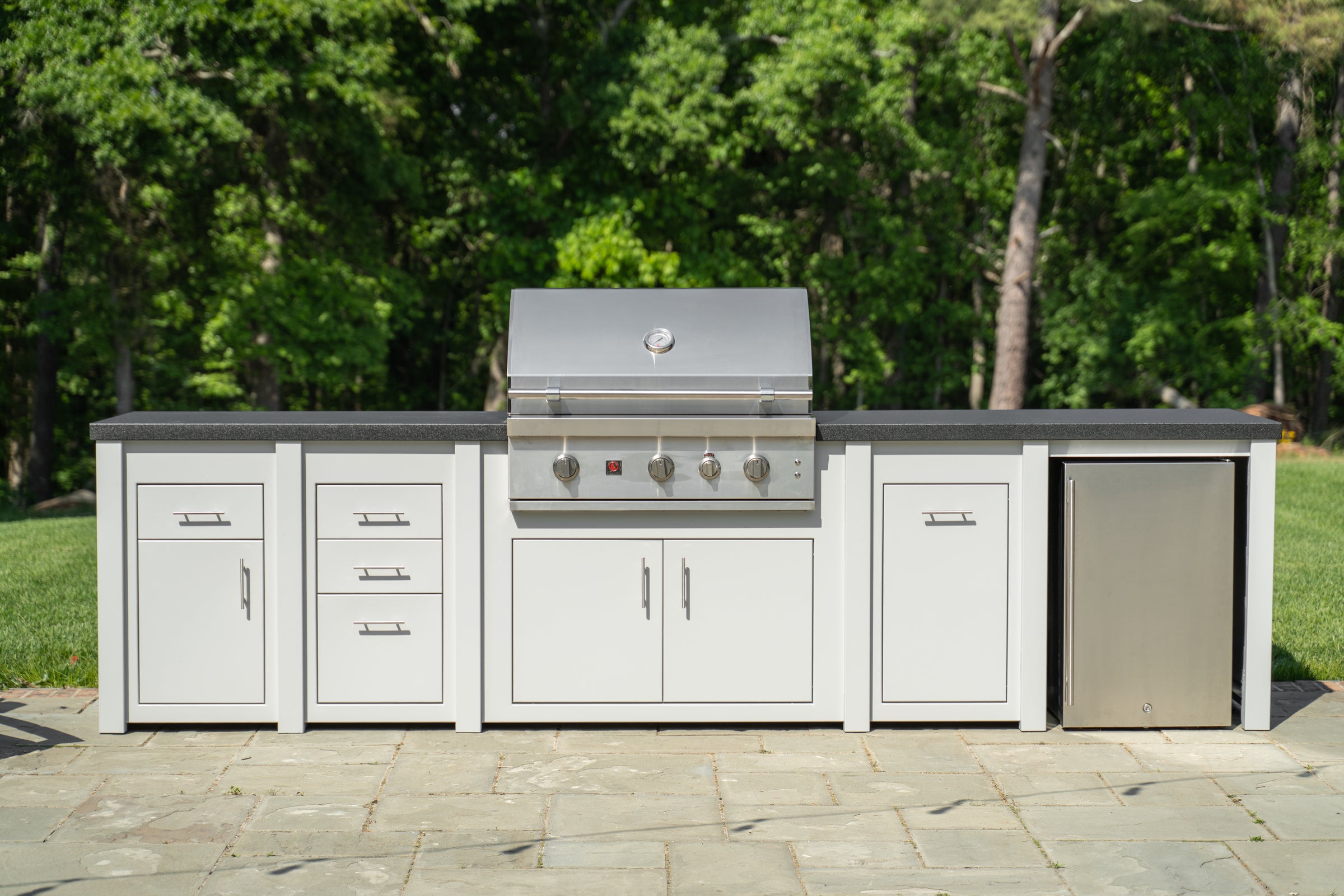 An outdoor kitchen island set with a stainless steel gas grill, white cabinets, a dark countertop, and a built-in refrigerator, set against a background of green trees and grass.