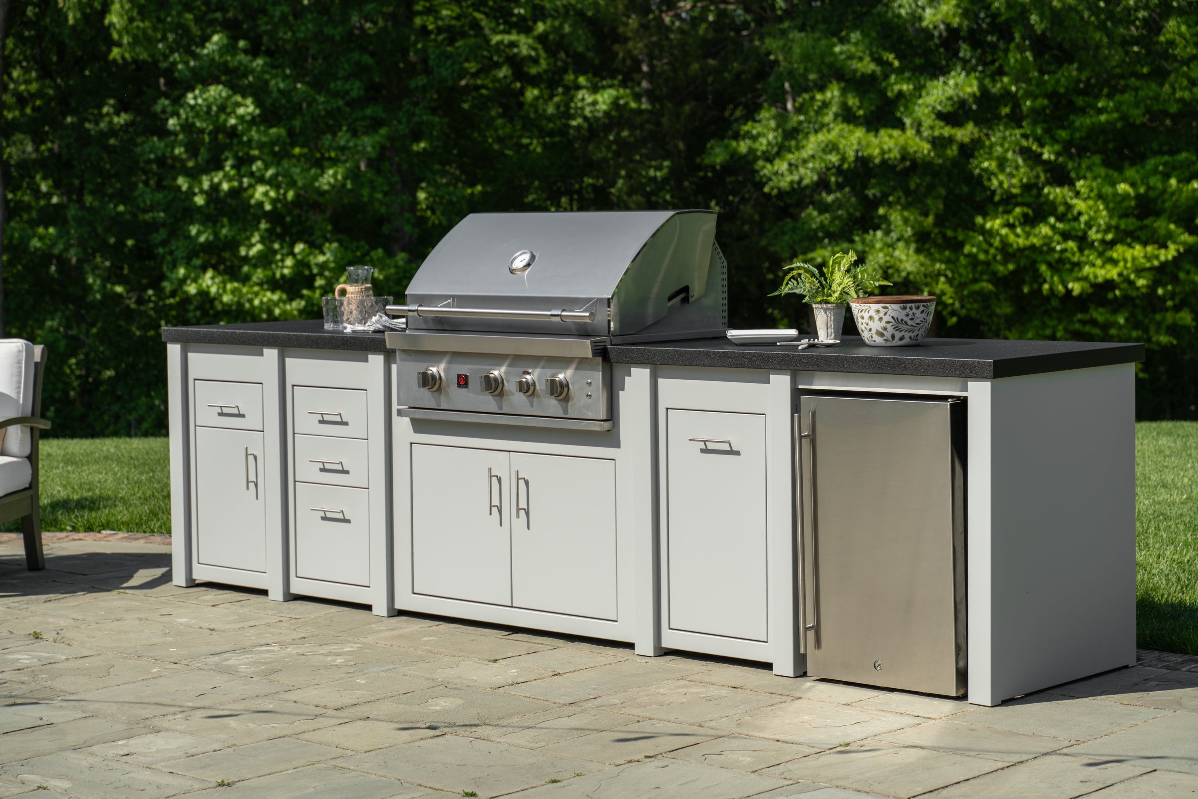 Outdoor kitchen island set with a stainless steel gas grill, white cabinets, a dark countertop, and a small stainless steel refrigerator, situated on a stone patio with green trees in the background.