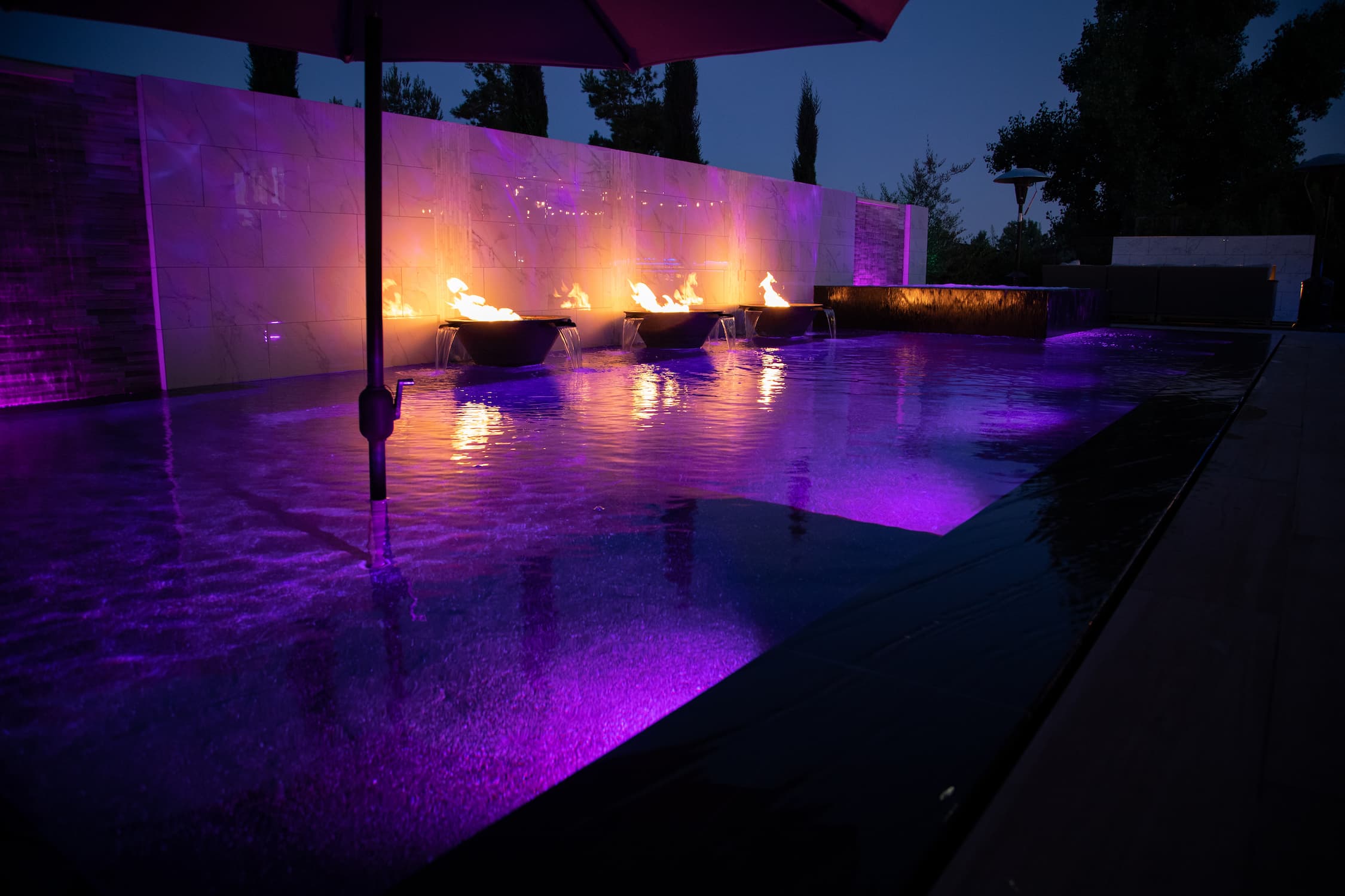 Modern outdoor pool area at dusk with purple and magenta water lighting, several lit fire pits, a reflective stone wall, and dark trees in the background.