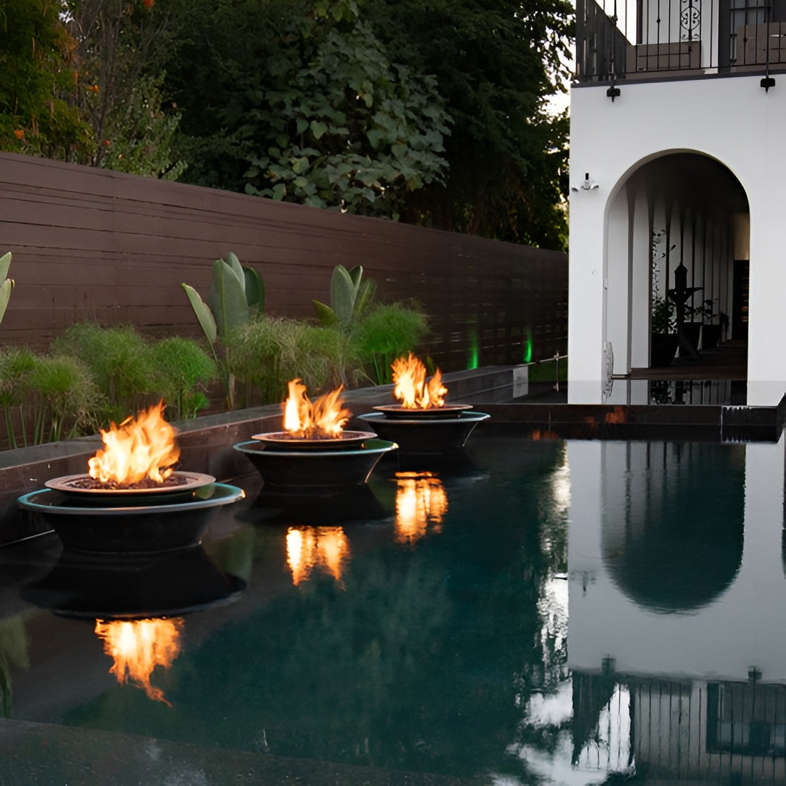 Three floating fire pits with bright flames sit in a rectangular swimming pool beside a white building with an arched entryway and dark wooden fencing.
